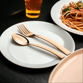 Gold spoon and fork placed on white plate beside pasta dish and drink on black surface.
